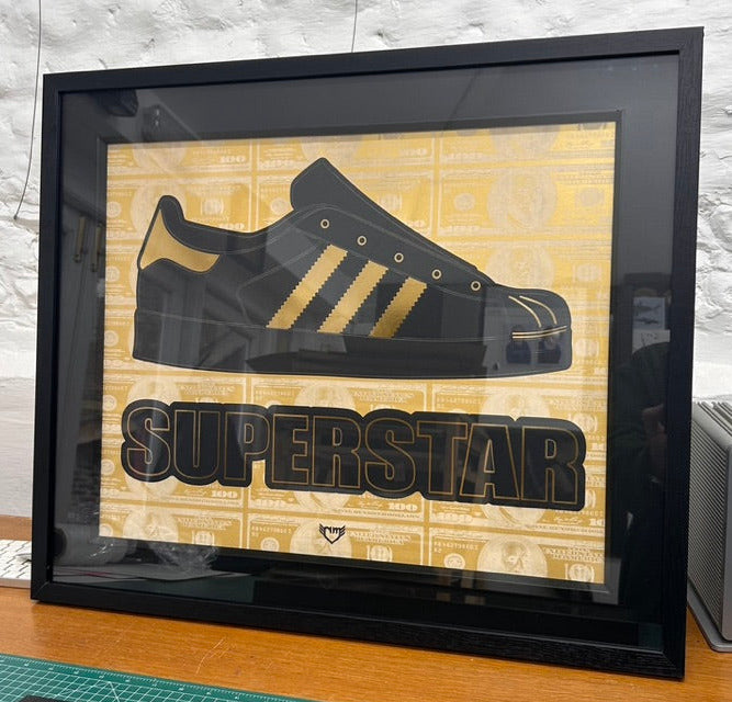 Framed picture of a black Adidas superstar shell toe shoe with gold stripes on a wooden surface.